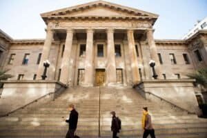 Three pedestrians walk past a large stone building with a grand staircase and six columns.