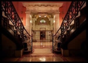 Ornate dark wood staircases with wrought iron railings flank a marble archway leading to a gated hallway with a chandelier.